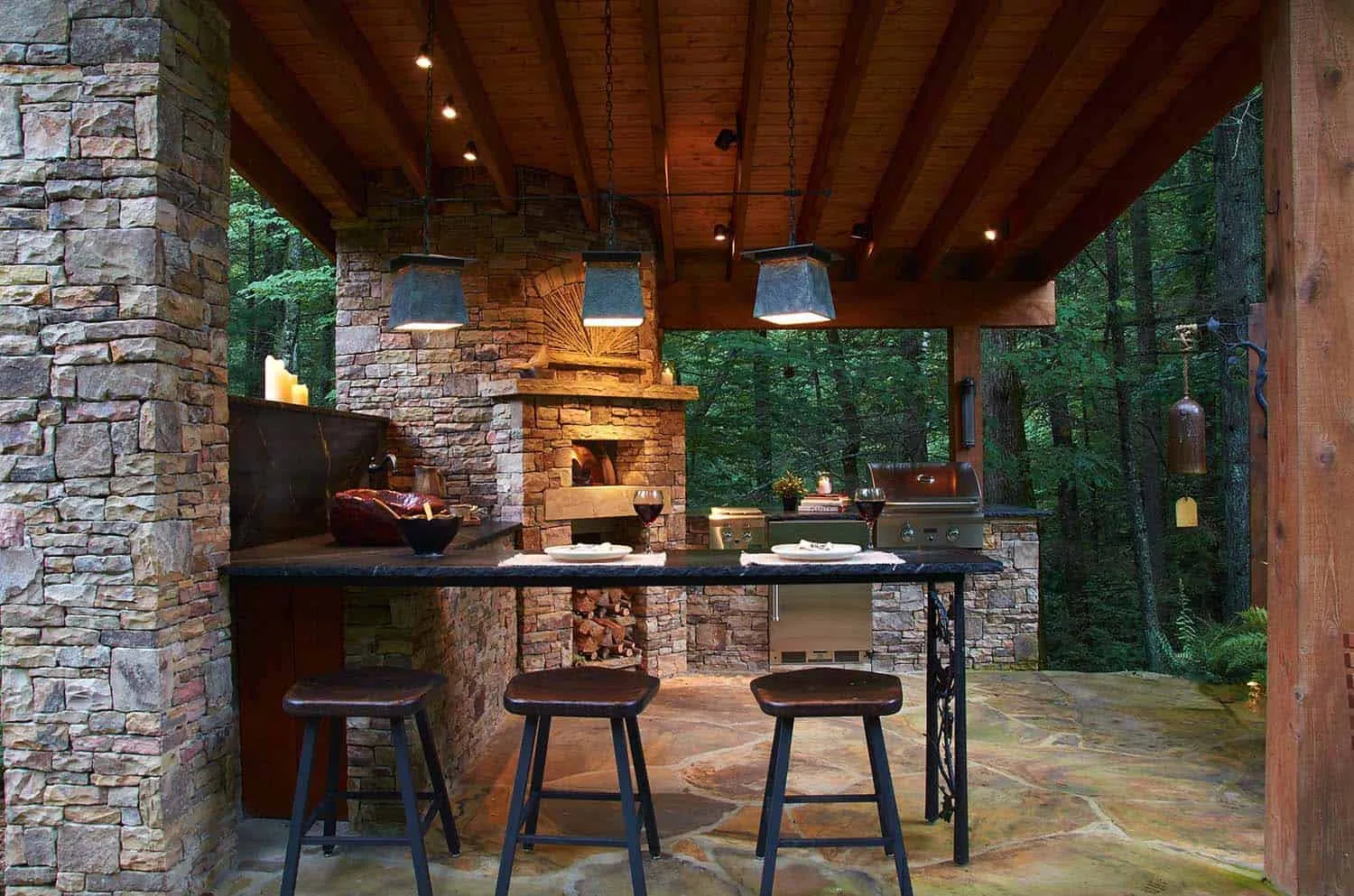 Luxurious outdoor kitchen with stone fireplace, bar, grill, and forest views.