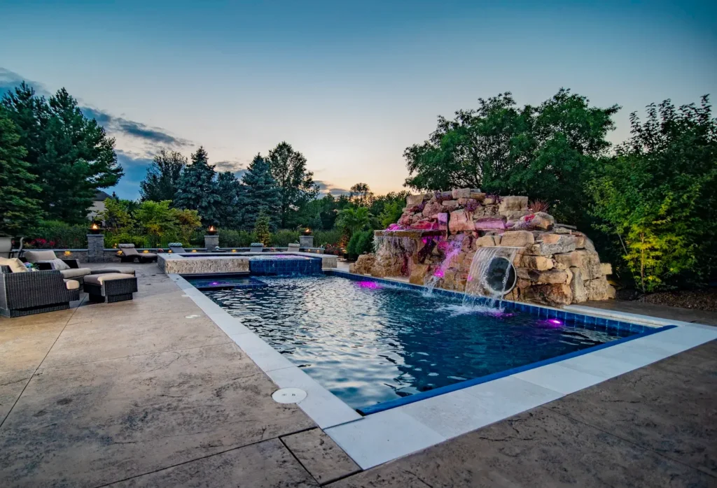 Illuminated swimming pool with rock waterfall, hot tub, and patio furniture at twilight.