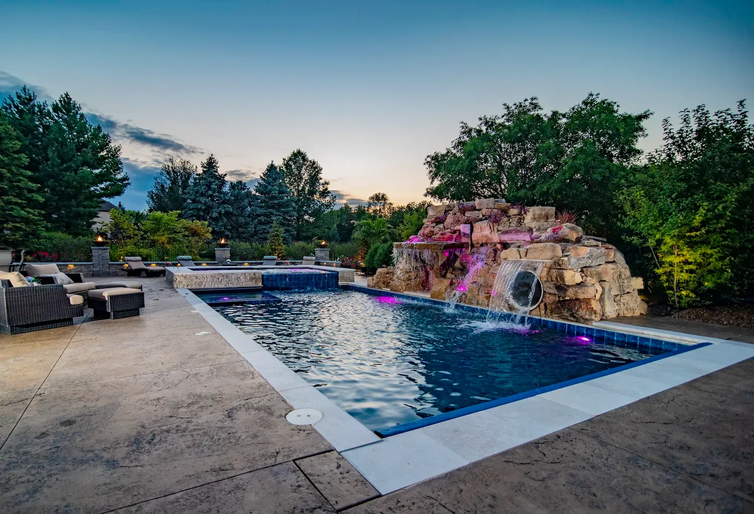 Illuminated swimming pool with rock waterfall, hot tub, and patio furniture at twilight.